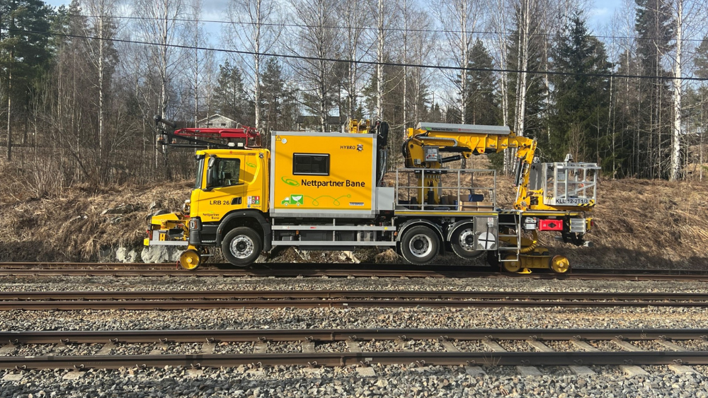 Sidevisning av hybrid stormobil fra Nettpartner Bane parkert på skinner med skog i bakgrunnen