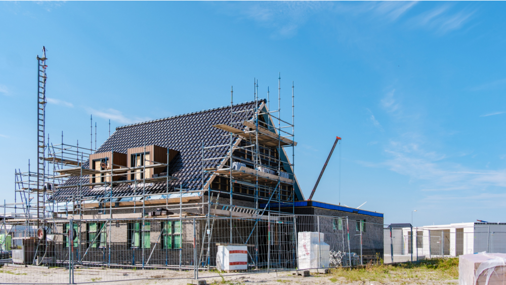 Et hus under oppføring med stillas, takarbeid og byggeplassgjerder, på en solrik dag med blå himmel.